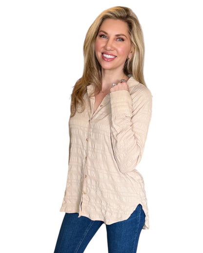 Blonde woman smiling while wearing the Almond Button Down Shirt paired with blue jeans. The shirt features a textured fabric, long sleeves, and a relaxed fit with a curved hem. 