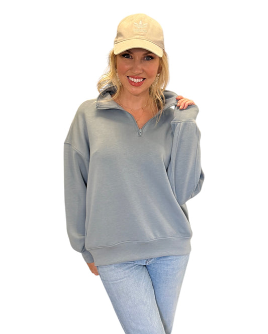 Woman smiling in jeans, cream baseball cap and Misty Blue Half-Zip Pullover with a relaxed fit, super-soft fabric, and hip-length hem, featuring a streamlined pocket-free design and adjustable half-zip neckline.