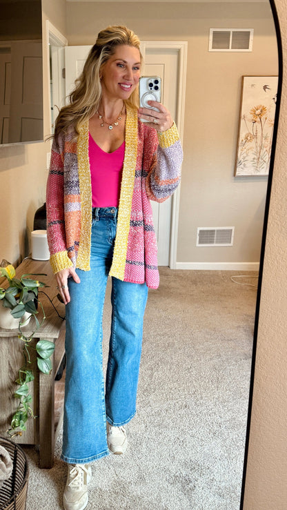 Woman taking a selfie in a mirror and wearing the Tenley Knit Dreamer Cardigan in vibrant purple, coral, and yellow tones, paired with a bright pink tank top and mid-rise wide-leg jeans. She accessorizes with a custom charm necklace made at Wish Upon Boutique. 