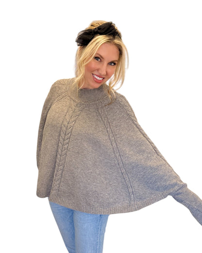 Woman wearing jeans and a Heather Grey/Taupe Cable Knit Mock Neck Poncho with a relaxed silhouette, cable knit design, and fitted sleeve hem, perfect for cozy layering.