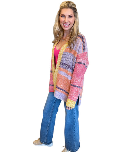 Woman wearing the Tenley Knit Dreamer Cardigan in vibrant purple, coral, and yellow tones, paired with a bright pink tank top and mid-rise wide-leg jeans. She accessorizes with a custom charm necklace made at Wish Upon Boutique, smiling against a white background.