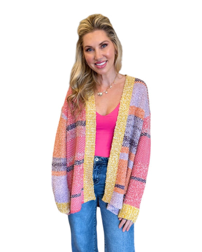 Woman wearing the Tenley Knit Dreamer Cardigan in vibrant purple, coral, and yellow tones, paired with a bright pink tank top and mid-rise wide-leg jeans. She accessorizes with a custom charm necklace made at Wish Upon Boutique, smiling against a white background.