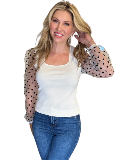 Woman wearing a white square-neck top with sheer, long polka dot sleeves, styled with blue jeans and soft waves in her blonde hair. 